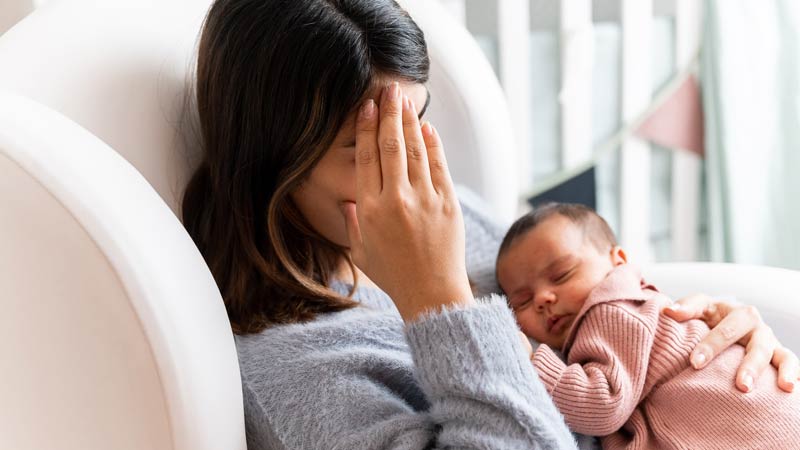 Mother sitting on couch in nursery feeling postpartum depression with baby in her arms.