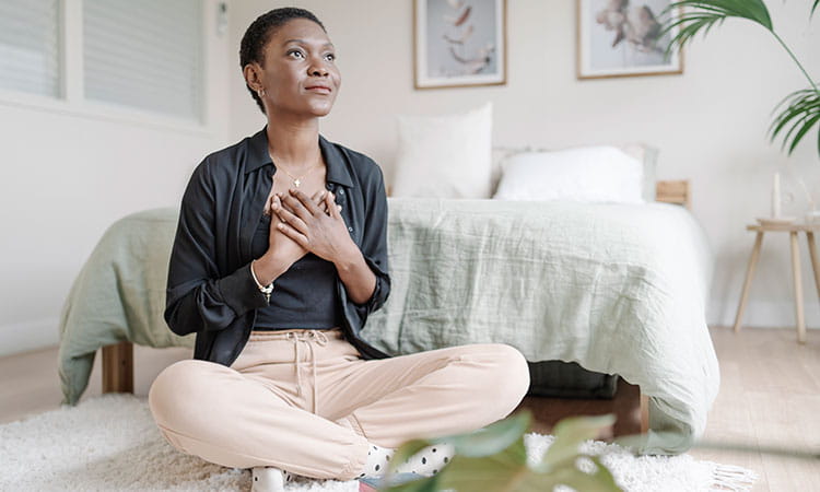 Mujer con las manos en el pecho meditando sentada en el suelo de su habitación.