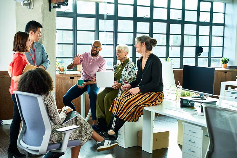 Grupo de empleados reunidos en la oficina, sentados en sillas y escritorios de manera casual