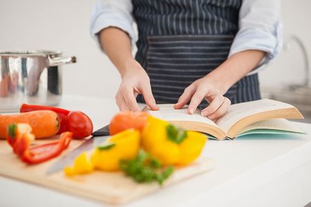 Manos que abren un libro de cocina, con pimientos morrones, zanahorias y una olla de fondo